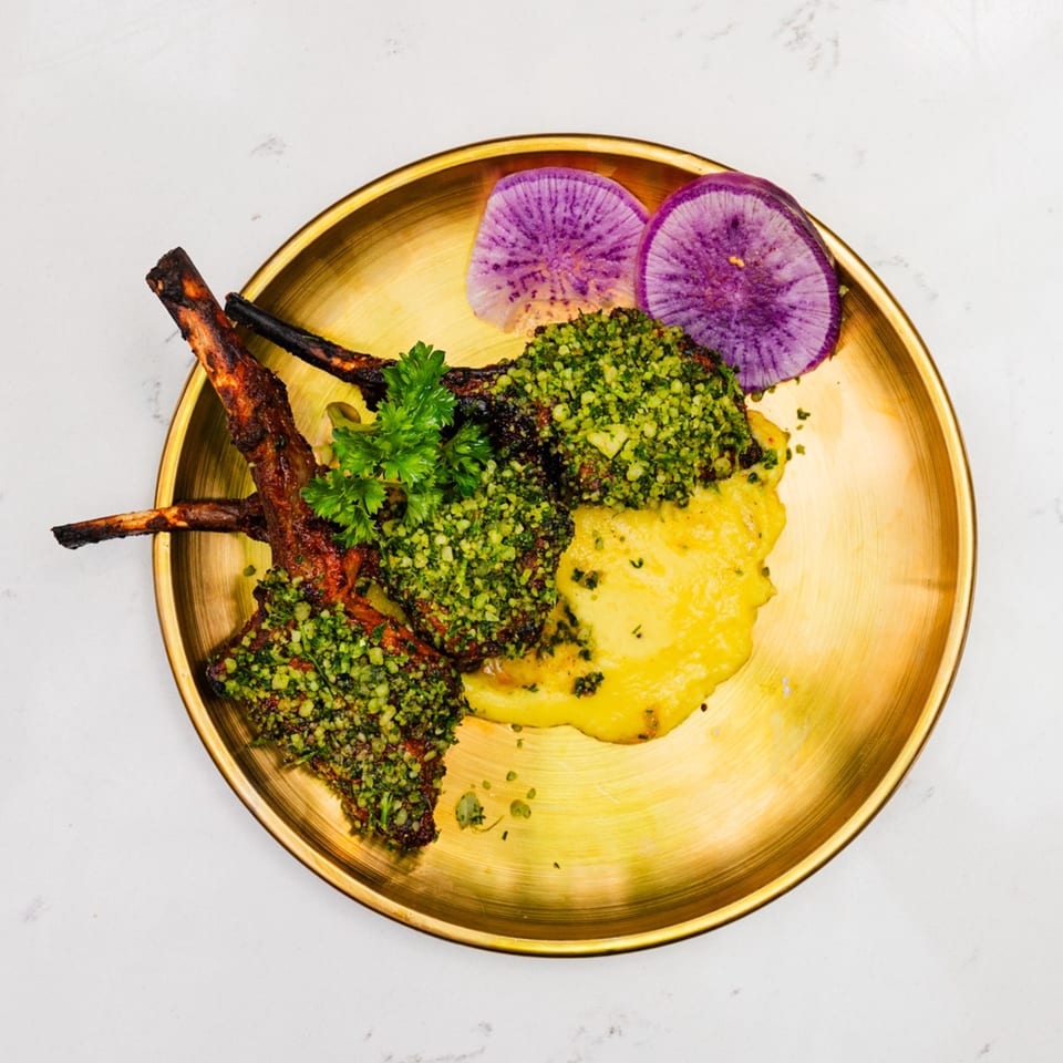 Indian lamb chops with green herb crust, yellow puree, and purple radish slices on a gold plate.