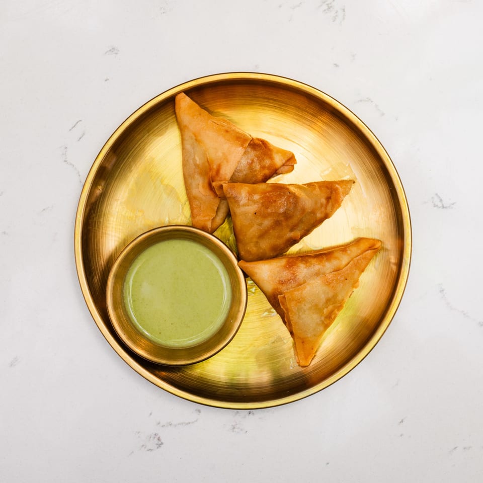 Golden brown samosas and a small bowl of green chutney on a brass plate.