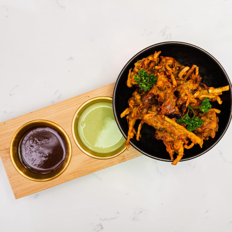 Crispy Indian onion bhaji in a black bowl with two dipping sauces on a wooden tray.