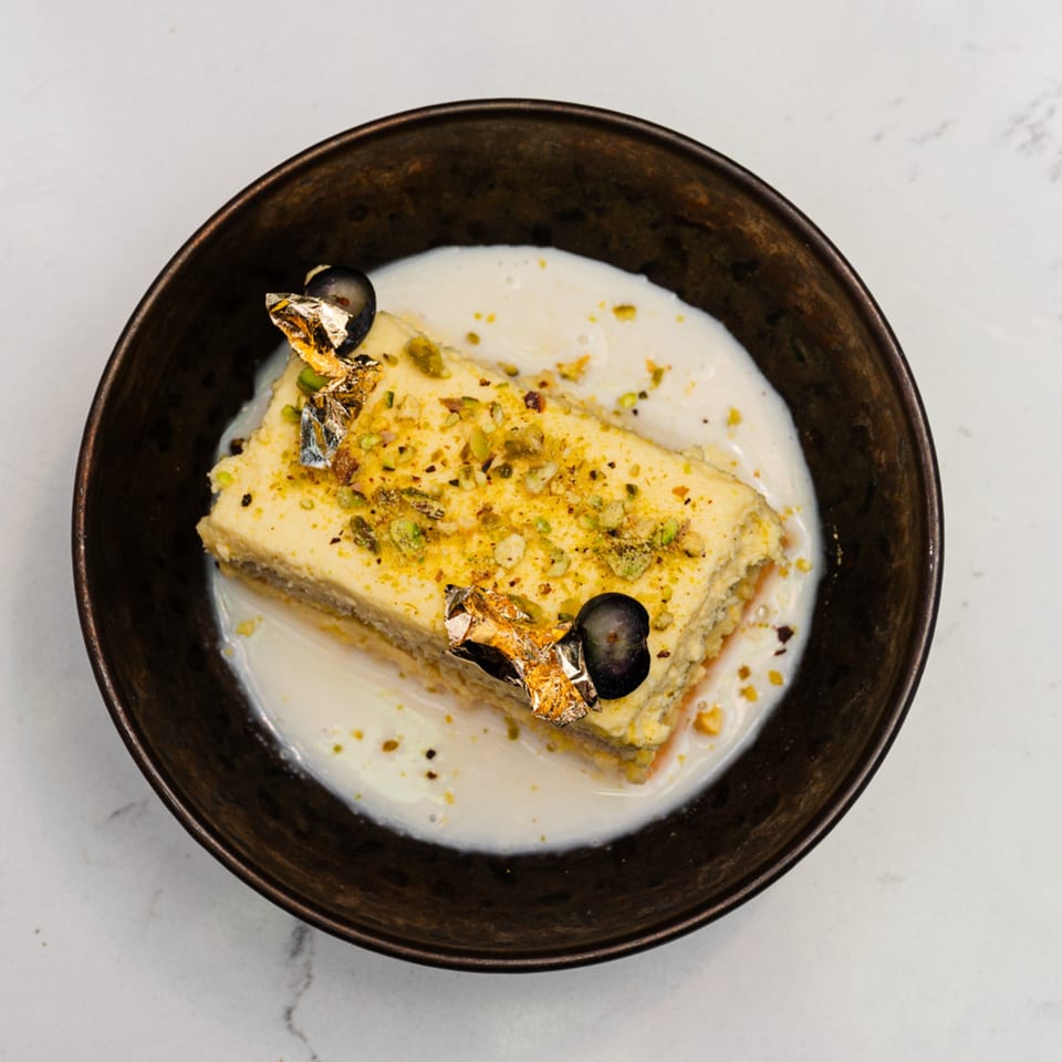 A rectangular Indian dessert, garnished with pistachios and gold leaf, sits in a dark bowl of cream.