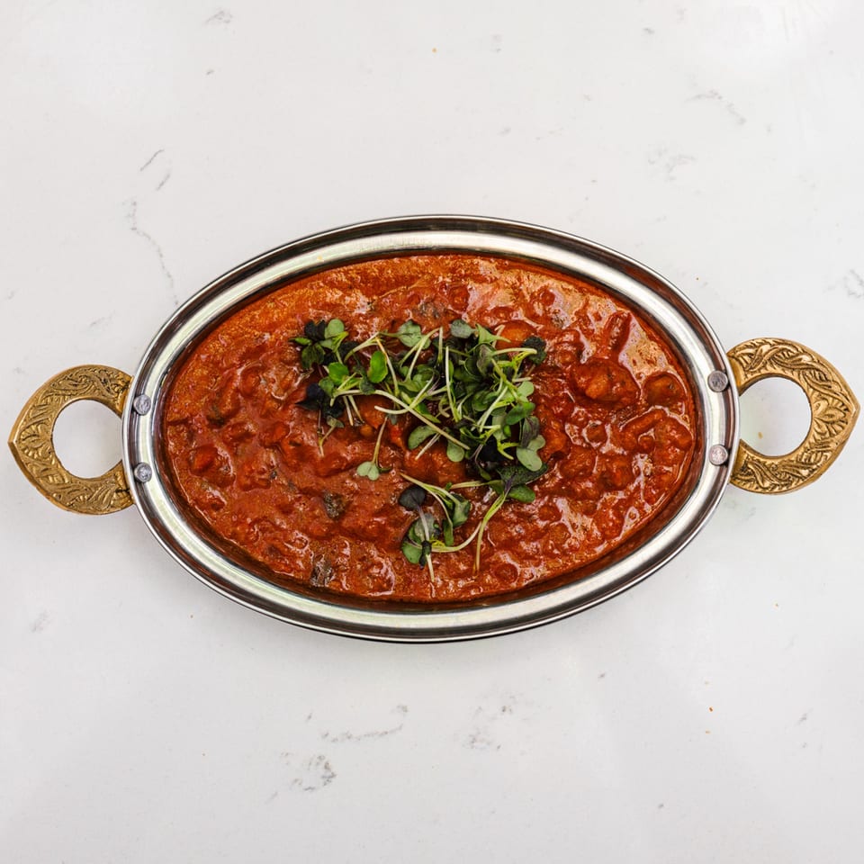 A vibrant red Indian curry, garnished with fresh microgreens, served in a silver oval dish.